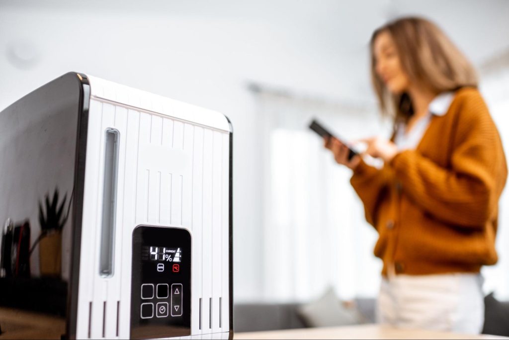 A woman remotely controls a smart air humidifier with her phone in a modern living room featuring a gray sofa and contemporary decor.