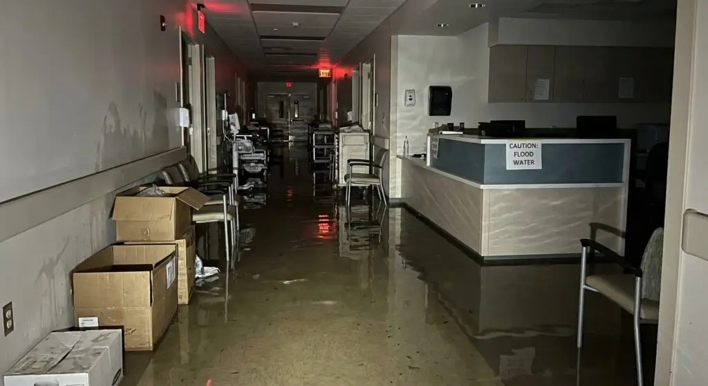 A dark, flooded hospital hallway with deep standing water, damaged medical supplies, and a "Caution: Flood Water" sign on a nursing station.