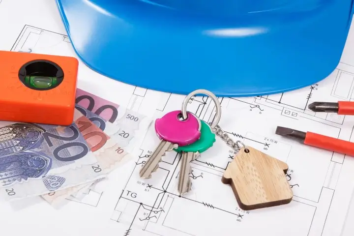 Keys with a house keychain beside a pile of cash on top of blueprints.