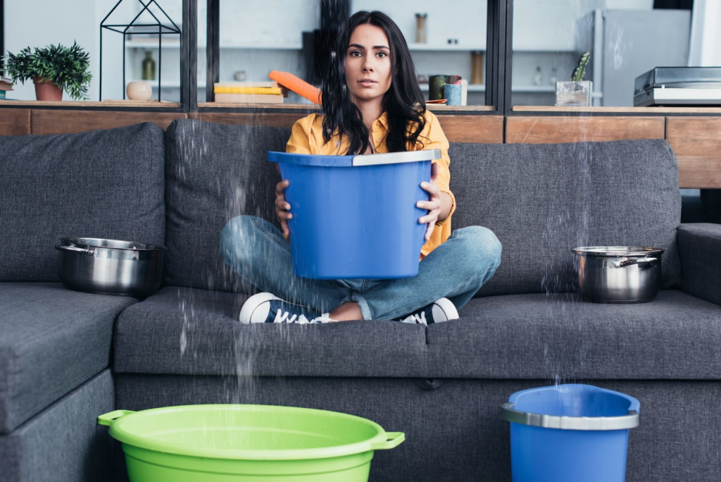 A woman holds a bucket over her couch and floors to prevent water damage.