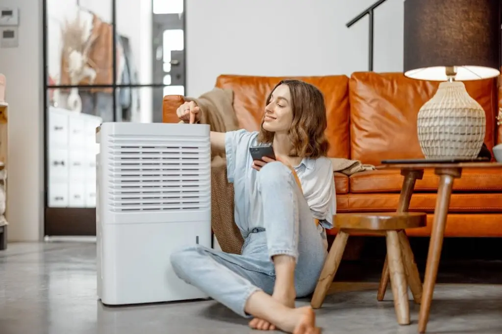A woman sits on the floor in front of an orange couch and turns on a dehumidifier.