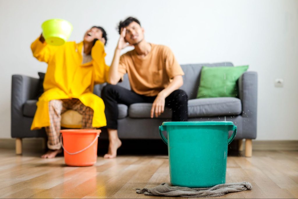 Two people sitting on a couch in a living room while water drips from the ceiling into buckets on the floor.