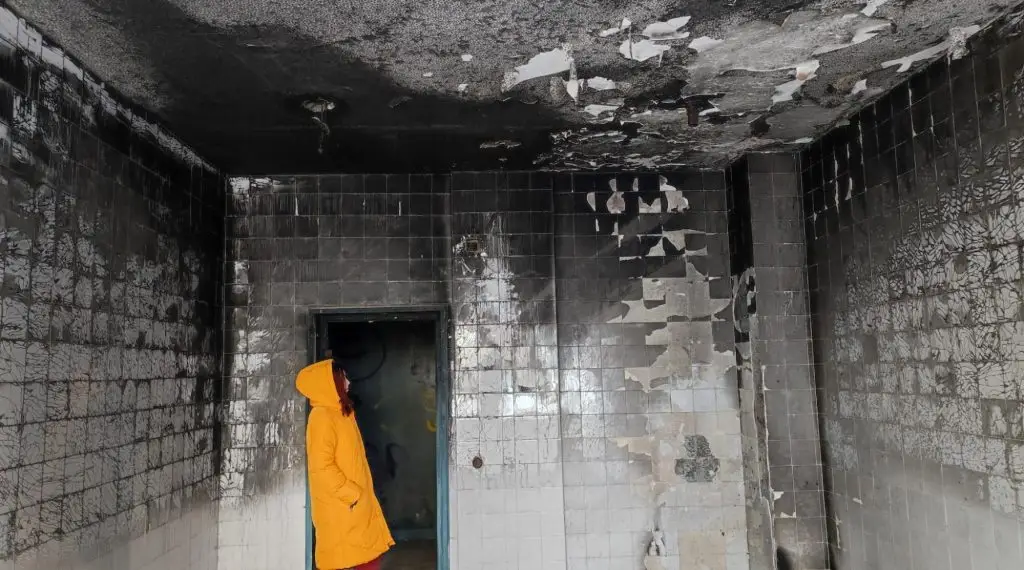 A young woman in a yellow hoodie stands at the doorway, surveying fire damage.