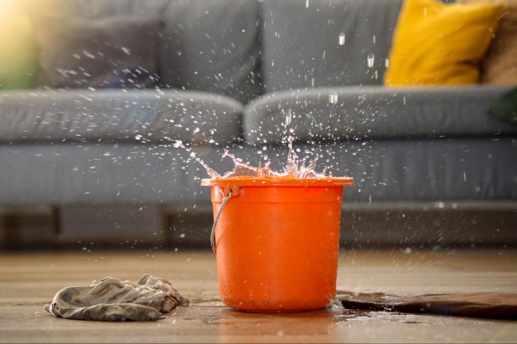 Water dripping from the ceiling into an orange bucket on the floor of a living room in front of a couch.