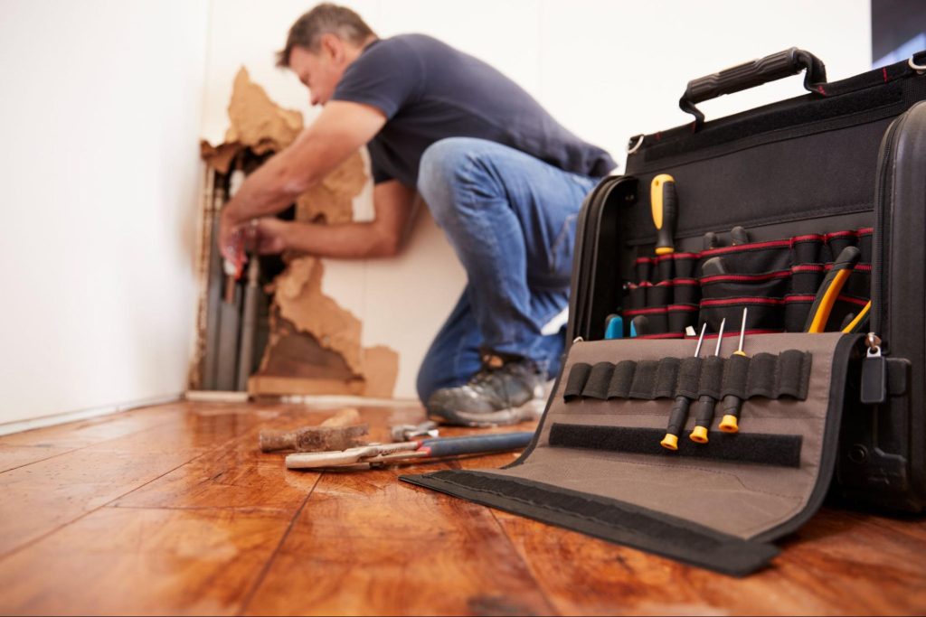A box of tools in the foreground with a worker in the background fixing a burst pipe that has damaged the wall.